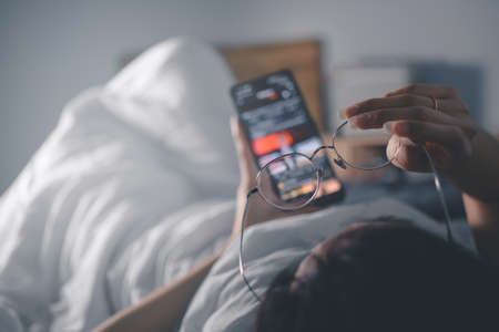Woman with glasses looks at smart phone and lies on bed, copy space for text.