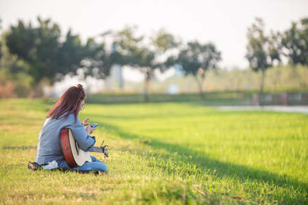 Young girl plays on her mobile phone while playing acoustic guitar in the park in the evening., copy space for text.の写真素材