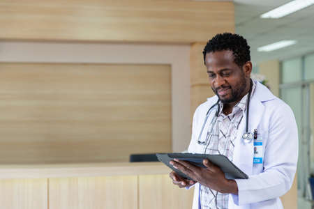 Male African doctor in white medical uniform fill patient medical history or anamnesis on tablet in hospital, man GP or physician consult client online on pad in modern clinic, healthcare conceptの写真素材