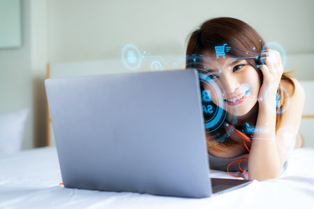 Beautiful Asian woman relaxing in bed while using a laptop to shop online on a sale day.の写真素材