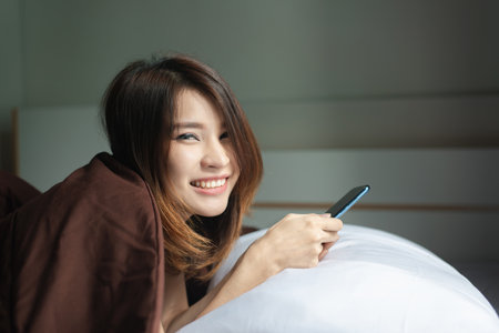 Attractive young Asian woman using phone in bedroom in the morningの写真素材