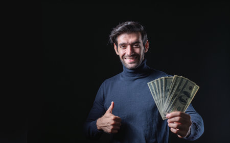 Portrait of a happy young businessman in a sweater displaying cash and giving the thumbs up, isolated on a black background with copy space for content.の写真素材