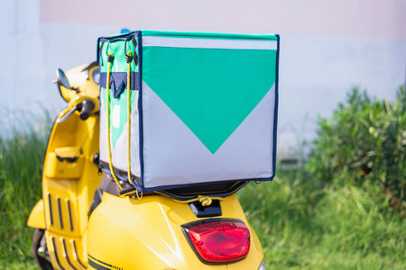 Yellow delivery scooter equipped with a large insulated food bag, parked outdoors with greenery in...
