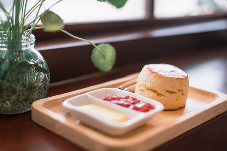 A freshly baked scone served with butter and strawberry jam on a wooden tray, next to a potted plant.の写真素材