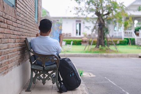 A man sits on a rustic bench holding a map, with a large backpack beside him, planning his travel outdoors in a peaceful setting.の写真素材