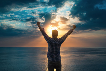 A man in a plaid shirt raises his arms triumphantly while standing by the ocean at sunrise under...