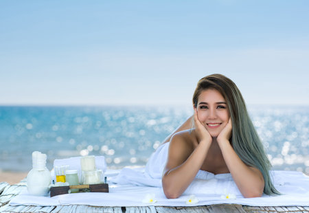 A cheerful woman enjoys a spa treatment on a sunny beach, surrounded by skincare products and the ocean's tranquil view.の写真素材