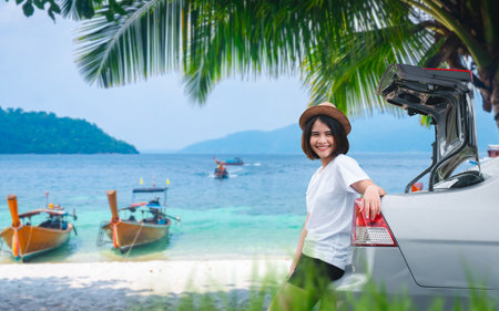 Happy young woman leaning on car trunk near tropical beach with wooden boats, palm trees, and blue ocean in the background.の写真素材