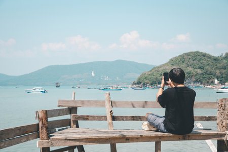 A man in casual clothing sits on a wooden bench by the sea, capturing the scenic coastal landscape with his smartphone.の写真素材