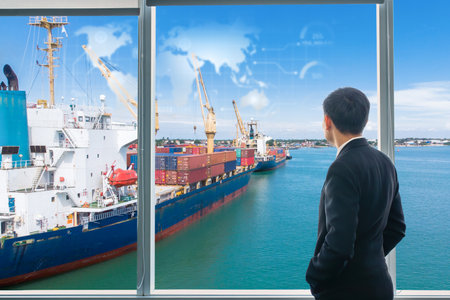 A businessman in a suit observes cargo ships loaded with shipping containers at a busy port,...