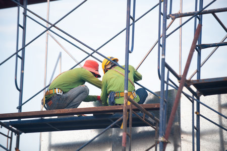 Two construction workers wearing safety gear work on metal structure while standing on scaffolding at a building site.の写真素材