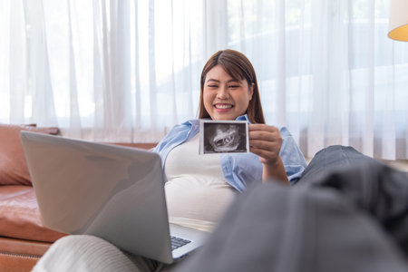Smiling pregnant woman relaxing on couch, holding baby ultrasound image while using laptop, sharing...