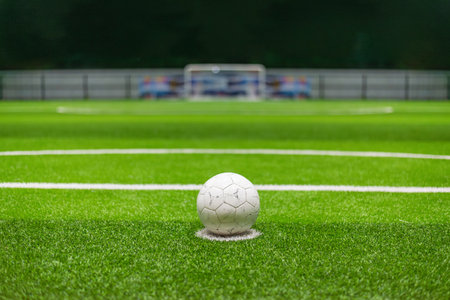 White soccer ball positioned on the center mark of a green football field, with goalposts in the background and illuminated stadium lighting, representing the start of a match or penalty shootout.の写真素材