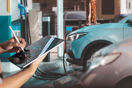 Technician using digital tablet for diagnostics and data analysis while an electric vehicle charges at a modern EV charging station. Concept of smart maintenance, clean energy, automotive technology.の写真素材