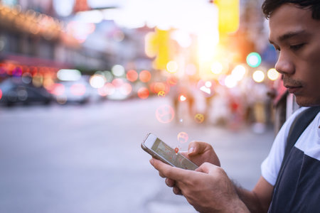 Young man holding smartphone and interacting with social media app, glowing emojis and location pin floating around, set against vibrant city street with bokeh lights during sunset.の写真素材