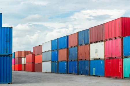 Stacks of multicolored shipping containers at a port storage yard under a cloudy sky, symbolizing...