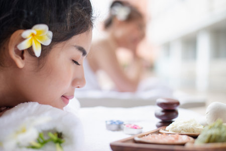 Close-up of a woman relaxing at a spa with a frangipani flower in her hair, surrounded by natural skincare products and massage tools.の写真素材