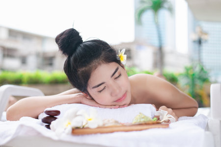 A woman lying on a massage bed with her eyes closed, with flowers and spa stones nearby. Represents wellness, beauty, and peaceful relaxation.の写真素材