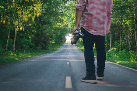 A person holding a camera stands on a quiet forest road, surrounded by lush green trees and morning sunlight ahead.の写真素材