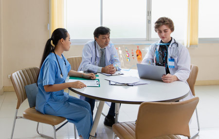 Doctors and a nurse collaborate around a table reviewing digital patient diagnostics on a laptop during a modern clinical briefing session.の写真素材
