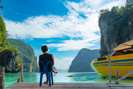 Back view of a man sitting on a chair on a wooden pier, looking at the beautiful turquoise sea, limestone cliffs, and a yellow speedboat. Tropical island vacation concept.の写真素材