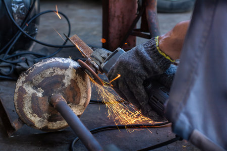 Close-up of industrial worker hands wearing gloves using an angle grinder to cut or polish rusty metal. Bright sparks flying in a workshop environment.の写真素材