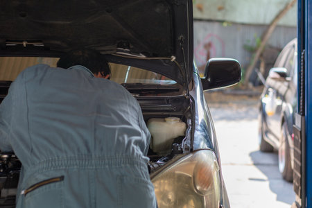 A professional auto mechanic in blue coveralls checking the engine under the open hood at a service station for vehicle maintenance and safety.の写真素材
