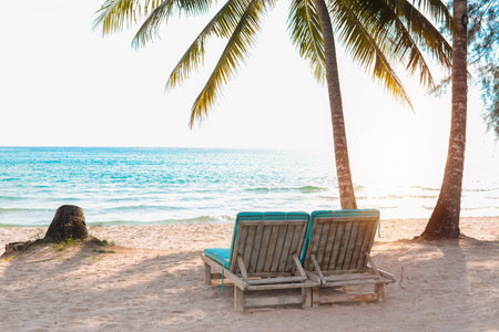 Two empty sun loungers on white sand beach with coconut palm trees and turquoise sea.の写真素材