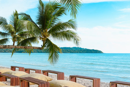 Relaxing tropical beach scene with wooden tables, beanbags, and a coconut palm tree overlooking the turquoise sea and island background.の写真素材