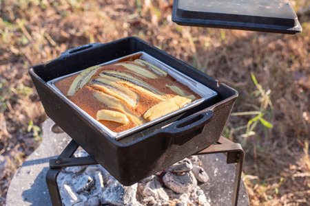 Freshly baked banana cake with banana slice toppings in a mold heating over hot charcoal embers. Concept of outdoor cooking, camping lifestyle, and homemade dessert.の写真素材