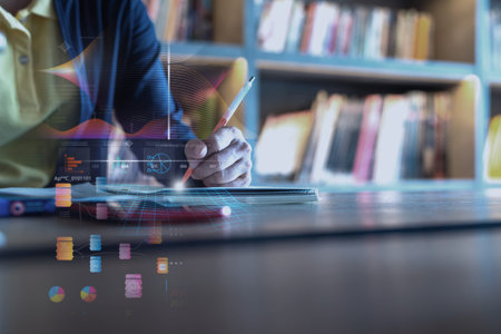 A person writing in a notebook at a desk with futuristic glowing digital data, business charts, and network interface overlays against a blurred library background.の写真素材