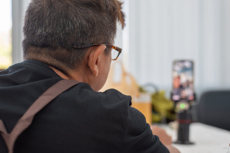 Rear view of a male entrepreneur live streaming to sell products online via smartphone. Concept of social commerce, digital marketing, and small business owner working from home.の写真素材