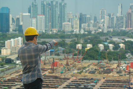 Back view of a male construction engineer in a yellow hard hat pointing at a massive urban construction site and city skyline.の写真素材