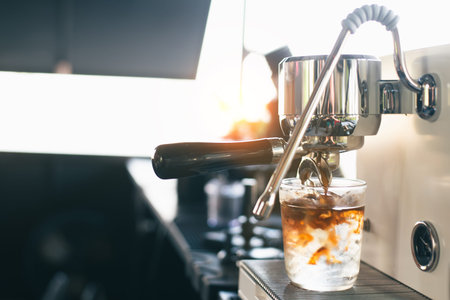 Close-up of hot espresso extracting from a portafilter directly into a glass of ice. Bright cafe setting with warm sunlight and beautiful coffee swirling effect.の写真素材