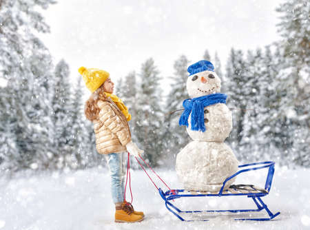 happy child girl plaing with a snowman on a snowy winter walk. child rolls a snowman on a sledの写真素材