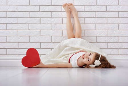 Sweet child girl with red heart. Happy little girl with Valentine's gift at a blank brick wall. Wedding, Valentine concept.の写真素材