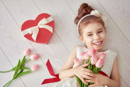 Sweet child girl with red heart and bouquet of tulips. Happy little girl with Valentine's gift lying on white wooden floor. Wedding, Valentine concept.の写真素材