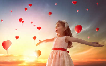 Sweet child girl looking at red balloons. Balloons in shape of heart flying in the sunset sky. Wedding, Valentine, love concept.の写真素材
