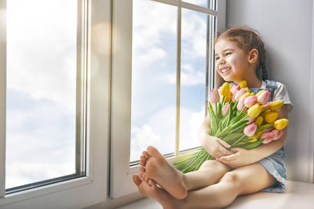 Adorable little child girl sitting on the window and holding tulips. Girl looks at the blue, pure sky and rejoices to spring and sun.の写真素材