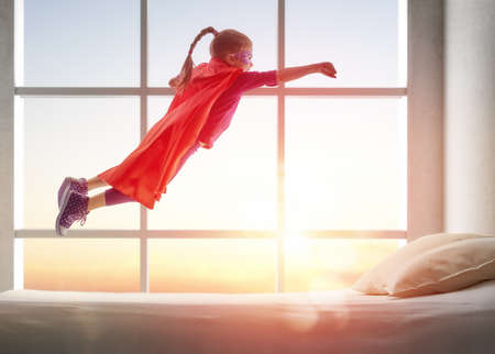 Child girl in Superhero's costume plays. The child having fun and jumping on the bed.の写真素材
