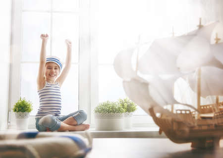 Adorable little child girl sitting on the window and holding lifeline. Girl looks at the blue sky and dreams of sea, adventures and travel. Girl has a toy ship.の写真素材
