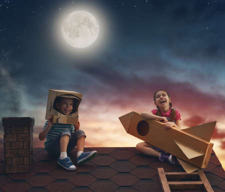 Two little children playing astronauts. Children on the background of moon sky. Child boy in an astronaut costume and child girl with toy rocket standing on the roof of the house and looking at the sky and dreaming of becoming a spacemen.の写真素材