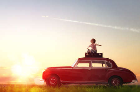 Toward adventure! Girl relaxing and enjoying road trip. Happy child girl sitting on roof of vintage car.の写真素材