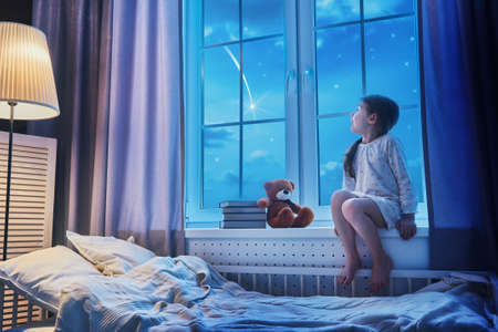 Cute child girl sitting at the window and looking at the stars. Girl making a wish by seeing a...