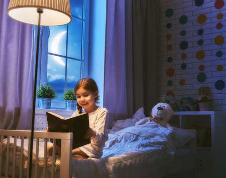 Cute little child girl reading a book under a lamp. Kid girl sitting in her bed on a dark moonlit night.の写真素材
