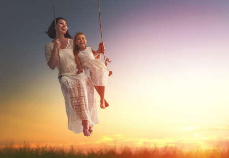Happy loving family! Young mother and her child daughter swinging on the swings and laughing a summer evening outdoors.の写真素材