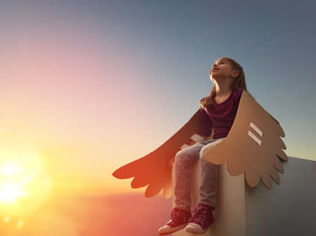 Little girl plays outdoors. Child on the background of sunset sky. Kid with the wings of a bird dreams of flying.の写真素材