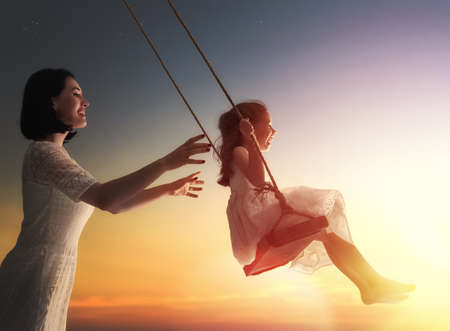 Happy loving family! Young mother and her child daughter swinging on the swings and laughing a summer evening outdoors.の写真素材