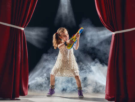 Cute little child girl playing guitar on stage. Kid dreams of becoming a rock musician.の写真素材