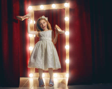 Cute little fashionista. Happy child girl try on outfits and mom's shoes looking at mirror.の写真素材